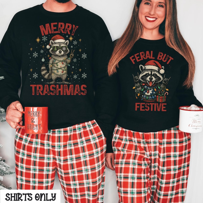 May include: Black sweatshirts with raccoon graphics and the text "MERRY TRASHMAS" and "FERAL BUT FESTIVE". The sweatshirts are paired with red and black plaid pajama pants. A red mug with "MERRY & BRIGHT" is visible.