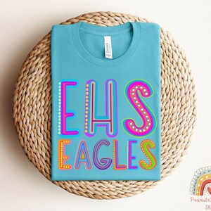 Puede incluir: Una camiseta azul claro con el texto "EHS EAGLES" en letras de colores y contorneadas. Las letras est&aacute;n decoradas con peque&ntilde;os puntos blancos.