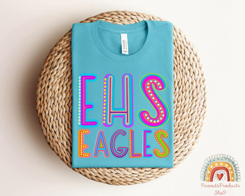Puede incluir: Una camiseta azul claro con el texto "EHS EAGLES" en letras de colores y contorneadas. Las letras est&aacute;n decoradas con peque&ntilde;os puntos blancos.