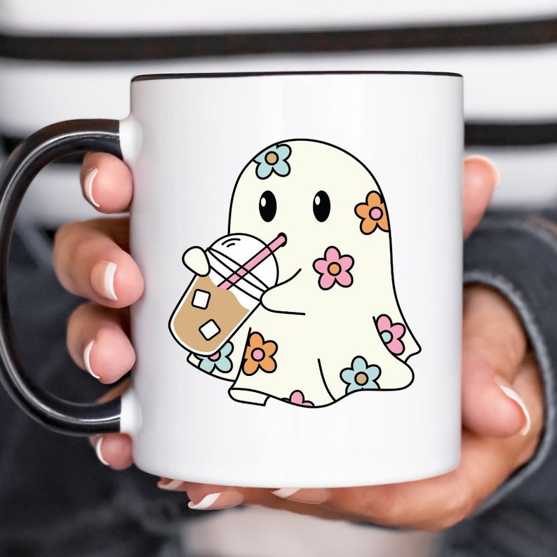 Op de afbeelding: Een witte keramische mok met een zwarte handgreep. De mok heeft een cartoon-geest met een bloemenpatroon die een kopje ijskoffie vasthoudt.