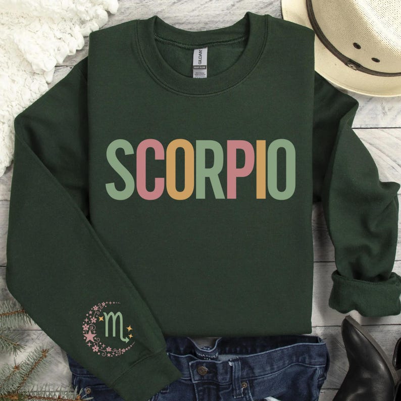 May include: A dark green sweatshirt with the word "SCORPIO" printed in rainbow colors. The sweatshirt has a crescent moon with a star and the Scorpio zodiac symbol printed on the left sleeve.