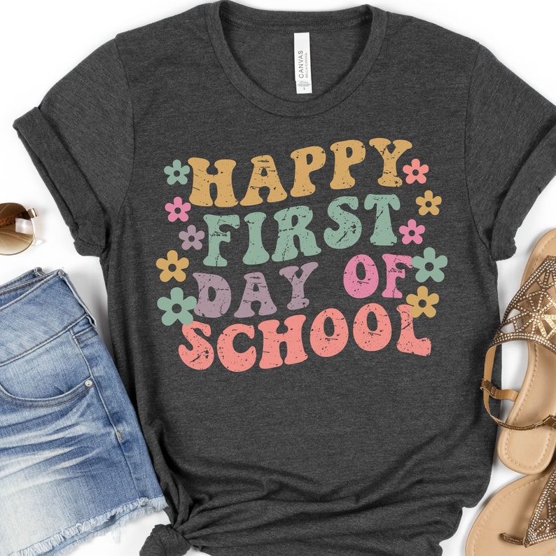 Puede incluir: Una camiseta gris oscuro con el texto "Happy First Day of School" en letras coloridas de estilo retro. El texto est&aacute; rodeado de peque&ntilde;as flores de colores.