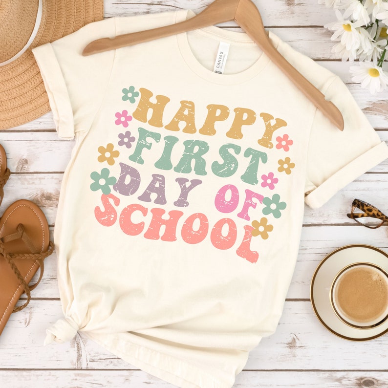 Puede incluir: Una camiseta blanca con el texto "Happy First Day of School" en una fuente retro con flores coloridas que rodean el texto.