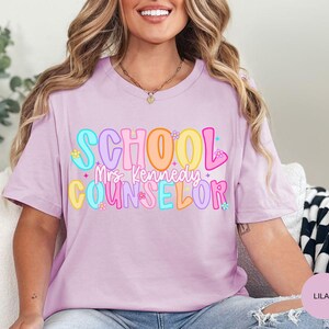 May include: A lilac-colored t-shirt with the words "SCHOOL Mrs. Kennedy COUNSELOR" in colorful bubble letters. The text is decorated with small flowers and stars. The shirt is a casual, everyday wear item.