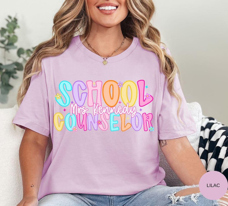 May include: A lilac-colored t-shirt with the words "SCHOOL Mrs. Kennedy COUNSELOR" in colorful bubble letters. The text is decorated with small flowers and stars. The shirt is a casual, everyday wear item.