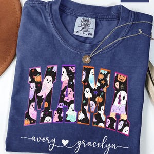 May include: A navy blue Comfort Colors t-shirt with the word "MAMA" in a colorful, spooky font filled with ghosts and pumpkins. The names "avery" and "gracelyn" are written below the word "MAMA". A silver necklace with a pendant is on the shirt.