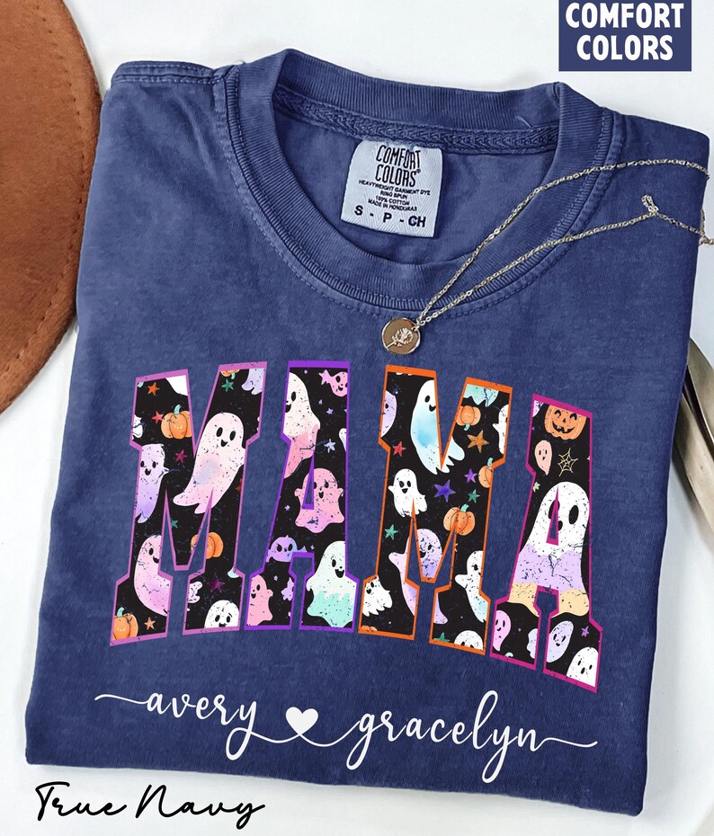 May include: A navy blue Comfort Colors t-shirt with the word "MAMA" in a colorful, spooky font filled with ghosts and pumpkins. The names "avery" and "gracelyn" are written below the word "MAMA". A silver necklace with a pendant is on the shirt.