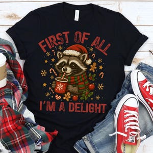 May include: Black t-shirt with a Christmas raccoon graphic. The raccoon wears a Santa hat and scarf, holding a cup of cocoa. Text reads "FIRST OF ALL I'M A DELIGHT" in red. Festive elements include candy canes, gingerbread men, and snowflakes. Red sneakers and jeans are visible.
