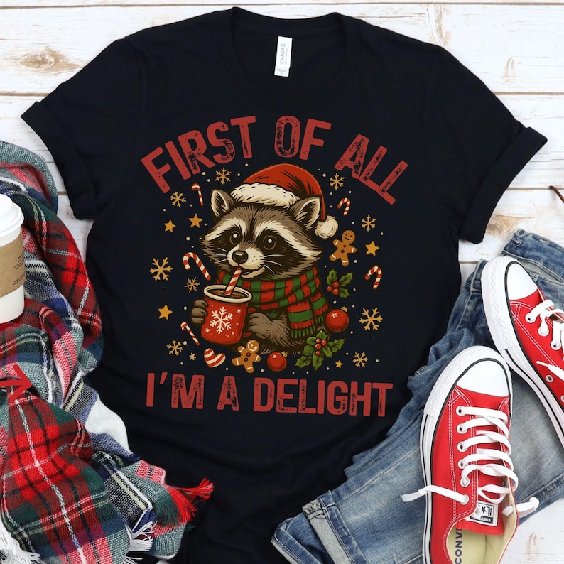 May include: Black t-shirt with a Christmas raccoon graphic. The raccoon wears a Santa hat and scarf, holding a cup of cocoa. Text reads "FIRST OF ALL I'M A DELIGHT" in red. Festive elements include candy canes, gingerbread men, and snowflakes. Red sneakers and jeans are visible.