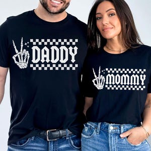 May include: Two black t-shirts with a white skeleton hand making a peace sign and checkered pattern. The text "Daddy" and "Mommy" is printed on the shirts.