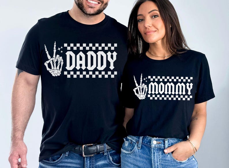 May include: Two black t-shirts with a white skeleton hand making a peace sign and checkered pattern. The text "Daddy" and "Mommy" is printed on the shirts.