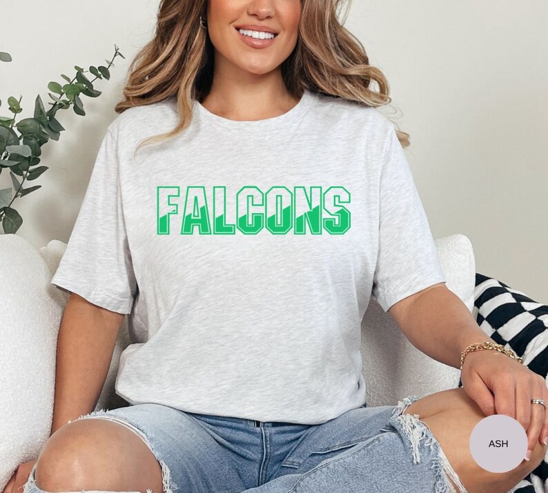 May include: A heather grey t-shirt with the word "FALCONS" printed in green letters. The letters have a white outline.