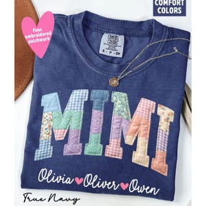 May include: A navy blue t-shirt with the word "MIMI" in patchwork letters. The patchwork includes floral, gingham, and solid fabrics in various colors. Below the name are the names Olivia, Oliver, and Owen. The shirt also has the text "True Navy" and "Comfort Colors".