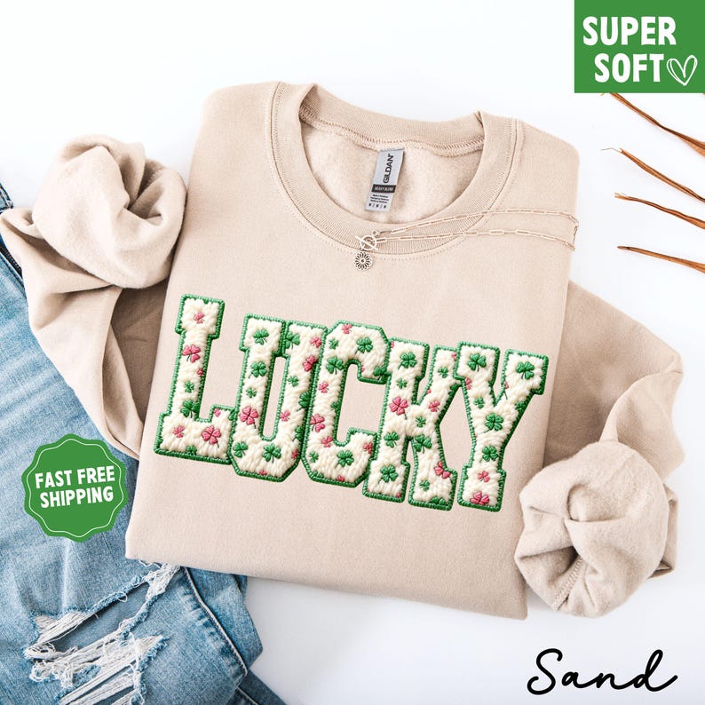 Pu&ograve; includere: Una felpa color sabbia con la scritta "LUCKY" ricamata in verde e motivi floreali. La felpa ha un girocollo e una collana d'argento. Sono visibili anche le parole "SUPER SOFT" e "Sand".