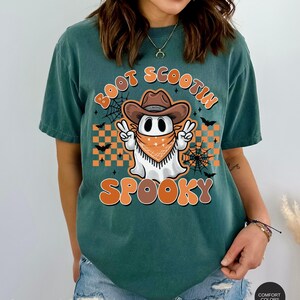 May include: A teal blue t-shirt with a graphic of a ghost wearing a cowboy hat and bandana. The text "Boot Scootin Spooky" is printed on the shirt.