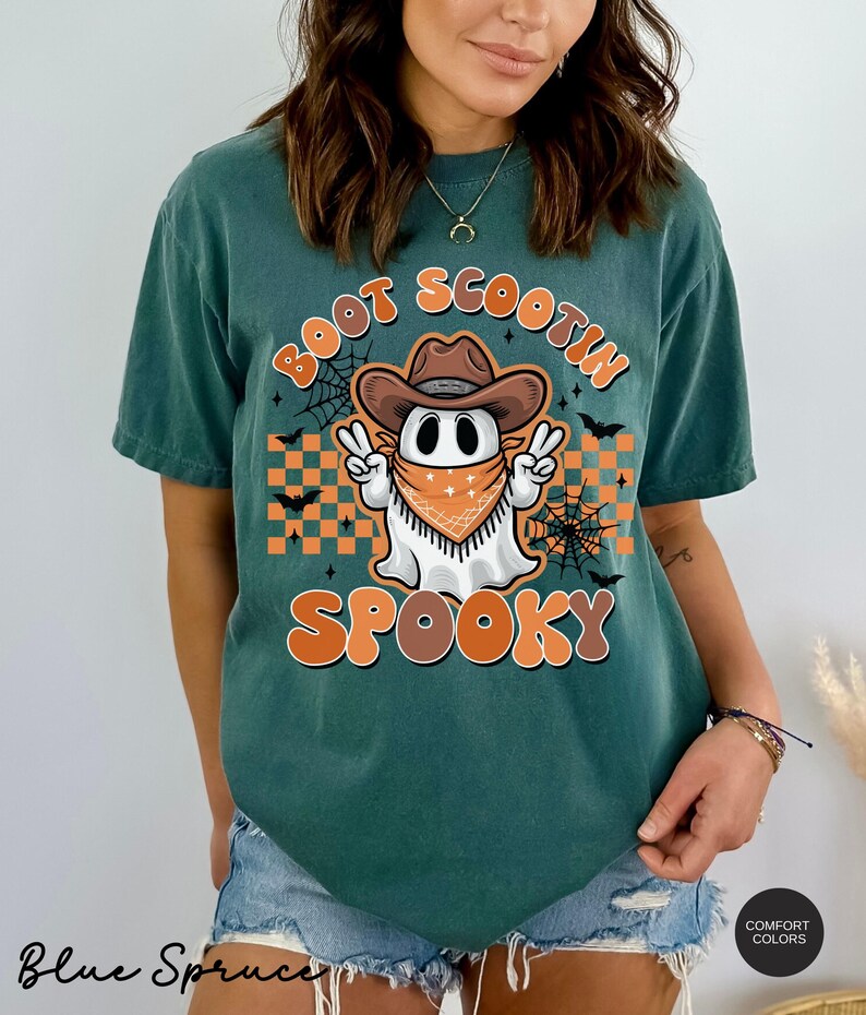 May include: A teal blue t-shirt with a graphic of a ghost wearing a cowboy hat and bandana. The text "Boot Scootin Spooky" is printed on the shirt.