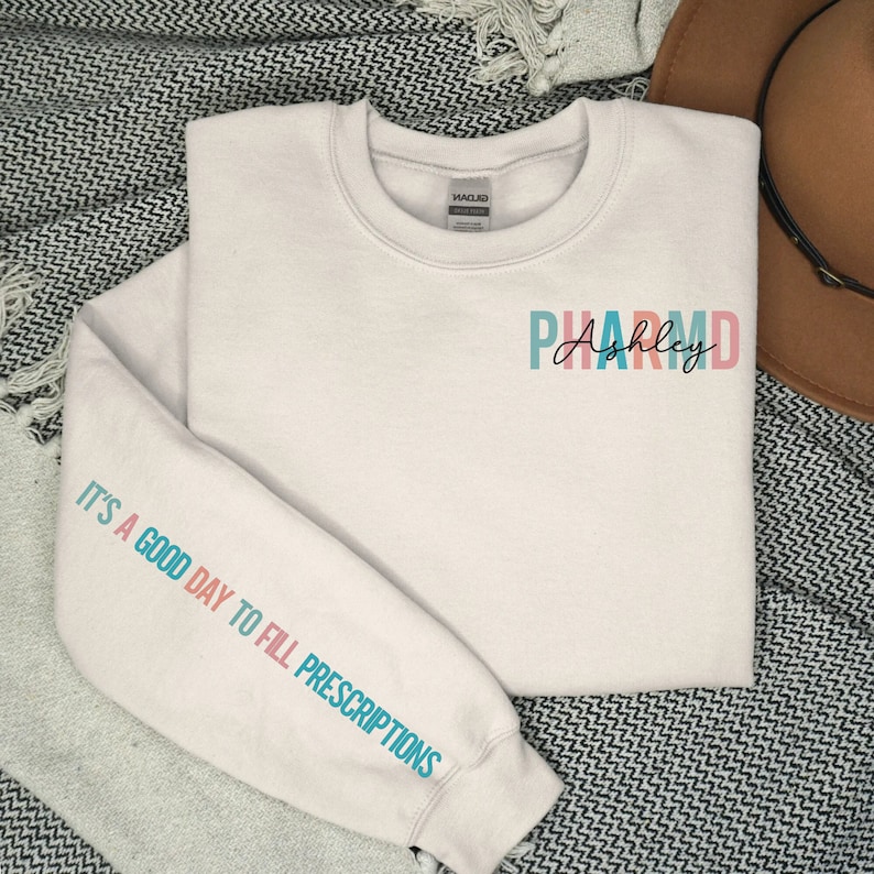 May include: White sweatshirt with the text "PHARM.D. Ashley" on the front and the text "It's a good day to fill prescriptions" on the sleeve.