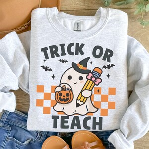 May include: A gray sweatshirt with a cute ghost wearing a witch hat and holding a pencil and a pumpkin. The text "Trick or Teach" is printed on the sweatshirt.