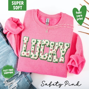 Pu&ograve; includere: Una felpa rosa con la scritta "LUCKY" ricamata in un motivo floreale verde e bianco. La felpa ha un girocollo e maniche lunghe. Il testo "Safety Pink" &egrave; scritto in corsivo in basso.