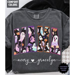 May include: Dark gray t-shirt with the word "MAMA" in colorful letters filled with ghost and pumpkin graphics. The names "avery" and "gracelyn" are written below the word "MAMA". The shirt has the text "CUSTOMIZE WITH ANY NAMES".