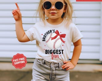 May include: A white t-shirt with a baseball graphic and the text "Brother's Biggest Fan". The shirt is decorated with a red bow and stars. The model is wearing the shirt and has blonde hair and is wearing sunglasses.