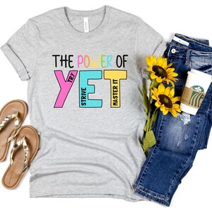 May include: A light grey t-shirt with the text "THE POWER OF YET" in colourful block letters. The shirt is paired with blue jeans, gold sandals, sunflowers, and a coffee cup.