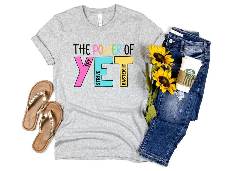 May include: A light grey t-shirt with the text "THE POWER OF YET" in colourful block letters. The shirt is paired with blue jeans, gold sandals, sunflowers, and a coffee cup.