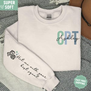May include: White sweatshirt with the text "CPT Ashley" in blue and green letters. The left sleeve has a dumbbell graphic and the text "Hit me with your best squat".