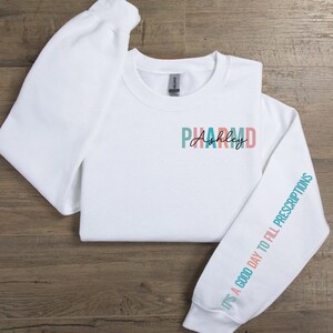 May include: White sweatshirt with the text "PHARM D Ashley" on the front and the text "It's a good day to fill prescriptions" on the sleeve in teal, pink, and peach colors.