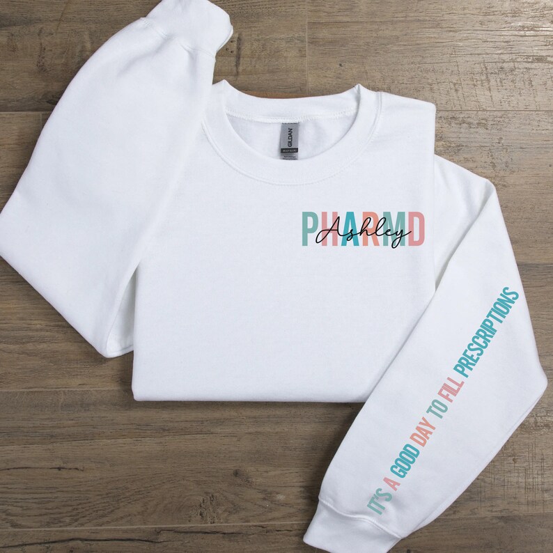 May include: White sweatshirt with the text "PHARM D Ashley" on the front and the text "It's a good day to fill prescriptions" on the sleeve in teal, pink, and peach colors.