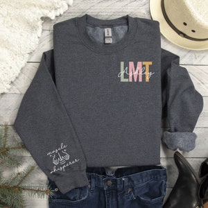 May include: A dark grey sweatshirt with the text "LMT Ashley" on the front and "muscle whisperer" on the sleeve. The sweatshirt is made of a soft, comfortable material and is perfect for everyday wear.