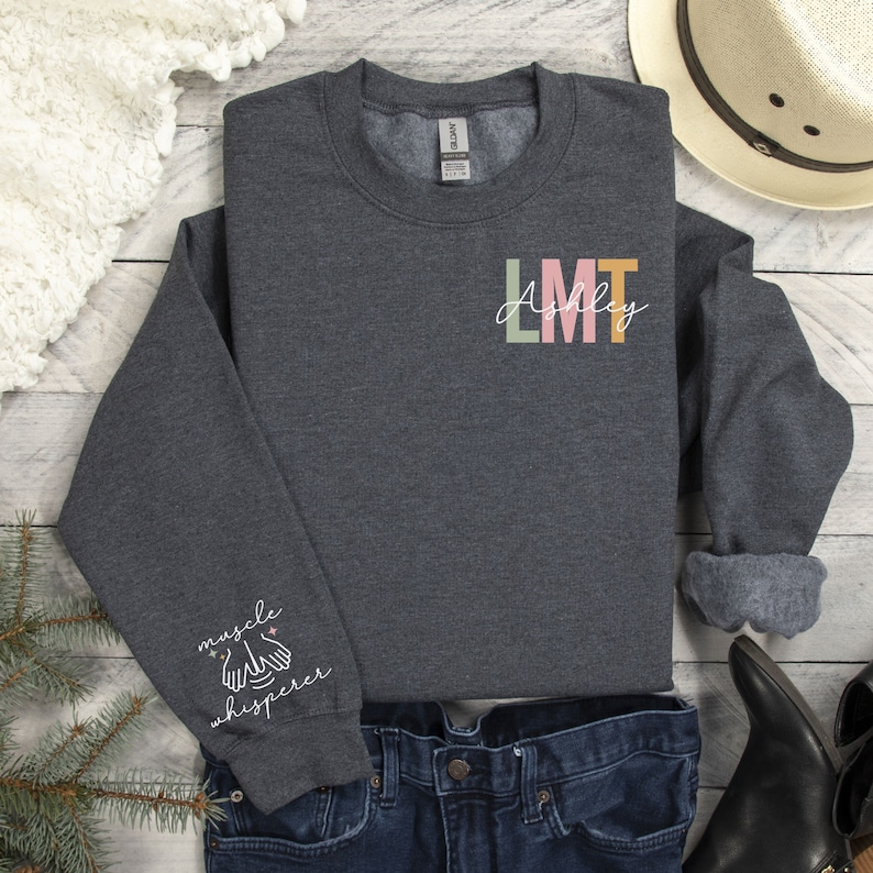 May include: A dark grey sweatshirt with the text "LMT Ashley" on the front and "muscle whisperer" on the sleeve. The sweatshirt is made of a soft, comfortable material and is perfect for everyday wear.