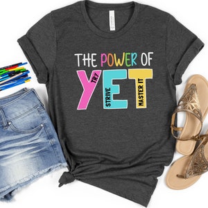 May include: Dark grey t-shirt with the text "THE POWER OF YET" in colourful block letters. The shirt is styled with denim shorts, sandals, and coloured pencils.