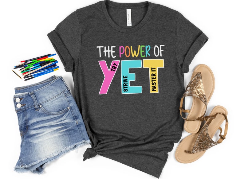 May include: Dark grey t-shirt with the text "THE POWER OF YET" in colourful block letters. The shirt is styled with denim shorts, sandals, and coloured pencils.