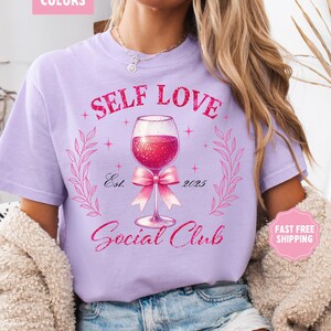 May include: A light purple t-shirt with the text "Self Love Social Club" in pink glitter lettering. A pink bow is tied around the stem of a glass of red wine with glitter. The text "Est. 2025" is below the wine glass.
