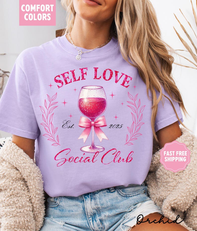 May include: A light purple t-shirt with the text "Self Love Social Club" in pink glitter lettering. A pink bow is tied around the stem of a glass of red wine with glitter. The text "Est. 2025" is below the wine glass.