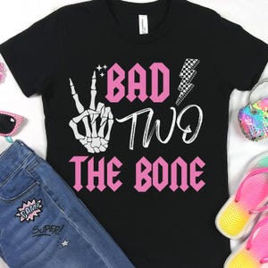 K&ouml;nnte beinhalten: Schwarzes T-Shirt mit einem pinkfarbenen Aufdruck, der "Bad Two The Bone" lautet. Der Aufdruck enth&auml;lt auch eine Skeletthand, die ein Peace-Zeichen macht, und einen Blitz.