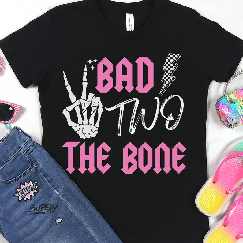 K&ouml;nnte beinhalten: Schwarzes T-Shirt mit einem pinkfarbenen Aufdruck, der "Bad Two The Bone" lautet. Der Aufdruck enth&auml;lt auch eine Skeletthand, die ein Peace-Zeichen macht, und einen Blitz.