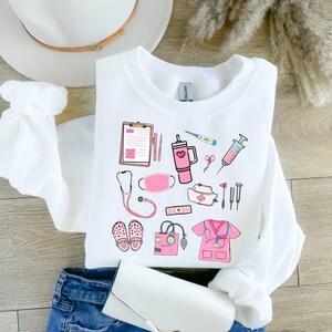 K&ouml;nnte beinhalten: Wei&szlig;es Sweatshirt mit einem rosa und schwarzen medizinischen Motiv. Das Design umfasst ein Stethoskop, eine Gesichtsmaske, einen Klemmbrett, ein Thermometer, eine Spritze und andere medizinische Werkzeuge. Das Sweatshirt ist auf einer blauen Jeans gefaltet.