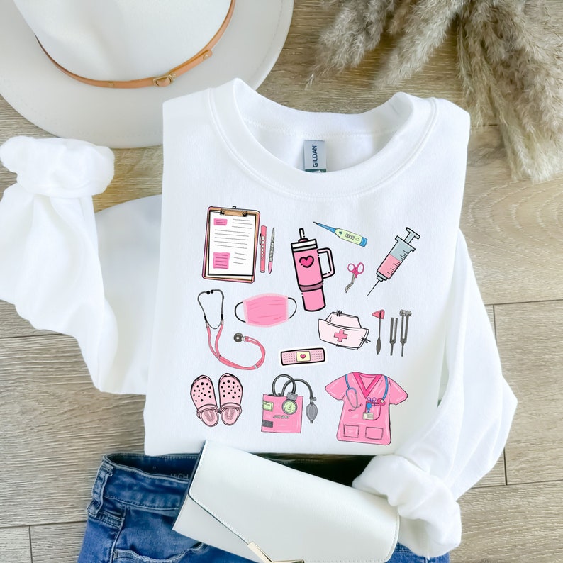 K&ouml;nnte beinhalten: Wei&szlig;es Sweatshirt mit einem rosa und schwarzen medizinischen Motiv. Das Design umfasst ein Stethoskop, eine Gesichtsmaske, einen Klemmbrett, ein Thermometer, eine Spritze und andere medizinische Werkzeuge. Das Sweatshirt ist auf einer blauen Jeans gefaltet.