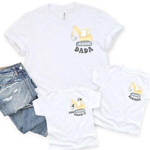 Peut inclure: T-shirts blancs avec des motifs d'excavatrice. L'un dit "DADA", un autre "BROTHER", et le troisi&egrave;me "I'M 2 AND DIGGIN IT". Un jean bleu est pli&eacute; &agrave; c&ocirc;t&eacute; des chemises. Les chemises sont faites d'une mati&egrave;re douce.