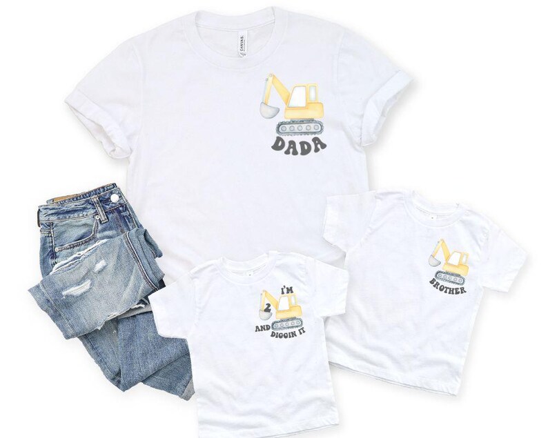 Peut inclure: T-shirts blancs avec des motifs d'excavatrice. L'un dit "DADA", un autre "BROTHER", et le troisi&egrave;me "I'M 2 AND DIGGIN IT". Un jean bleu est pli&eacute; &agrave; c&ocirc;t&eacute; des chemises. Les chemises sont faites d'une mati&egrave;re douce.