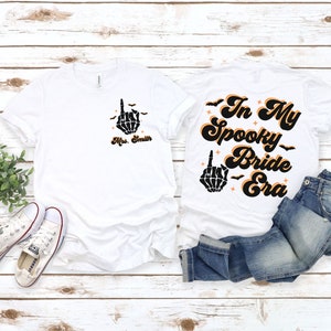 Puede incluir: Dos camisetas blancas con tem&aacute;tica de Halloween. Una camiseta dice "In My Spooky Bride Era" con una mano de esqueleto haciendo el dedo medio. La otra camiseta tiene una mano de esqueleto haciendo el dedo medio y el texto "Mrs. Smith".
