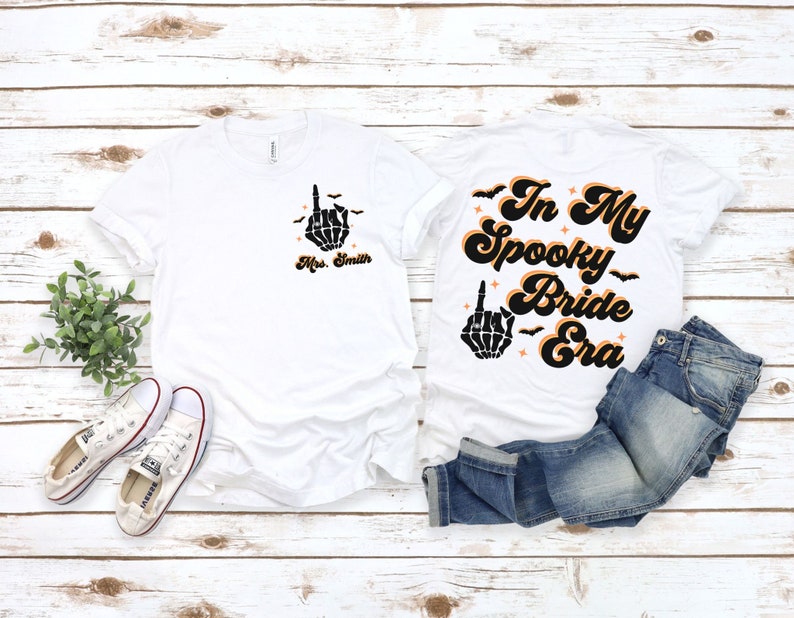 Puede incluir: Dos camisetas blancas con tem&aacute;tica de Halloween. Una camiseta dice "In My Spooky Bride Era" con una mano de esqueleto haciendo el dedo medio. La otra camiseta tiene una mano de esqueleto haciendo el dedo medio y el texto "Mrs. Smith".