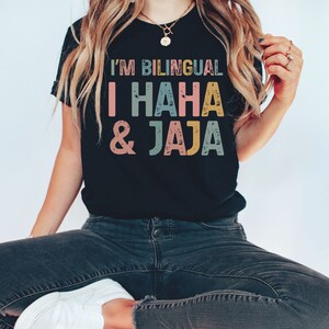 Puede incluir: Una camiseta negra con el texto "I'm Bilingual I HAHA & Jaja" en letras de colores desgastadas. El texto est&aacute; en una fuente de estilo vintage.