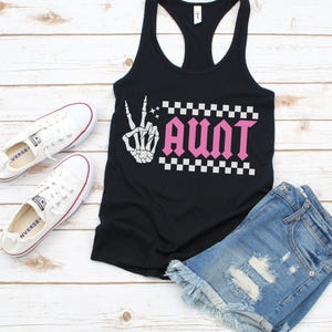 K&ouml;nnte beinhalten: Schwarzes Tanktop mit einer wei&szlig;en Skeletthand, die ein Peacezeichen macht, und dem Wort "AUNT" in rosa Buchstaben mit einem karierten Muster dahinter.