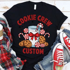 May include: Black t-shirt with a festive design. The graphic features the words "COOKIE CREW CUSTOM" in red, along with illustrations of cookies, candy canes, and a glass of milk tied with a red bow. Red sneakers and blue jeans are visible.