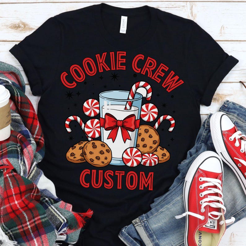 May include: Black t-shirt with a festive design. The graphic features the words "COOKIE CREW CUSTOM" in red, along with illustrations of cookies, candy canes, and a glass of milk tied with a red bow. Red sneakers and blue jeans are visible.