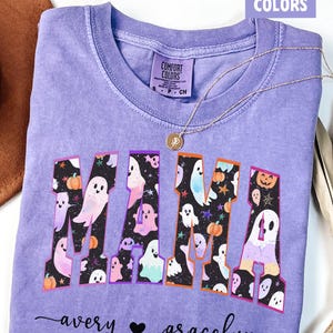 May include: A lavender Comfort Colors t-shirt with the word "MAMA" in a collegiate font, filled with ghost and pumpkin graphics. The names "avery" and "gracelyn" are written below the word "MAMA". A gold necklace with a pendant is on the shirt.