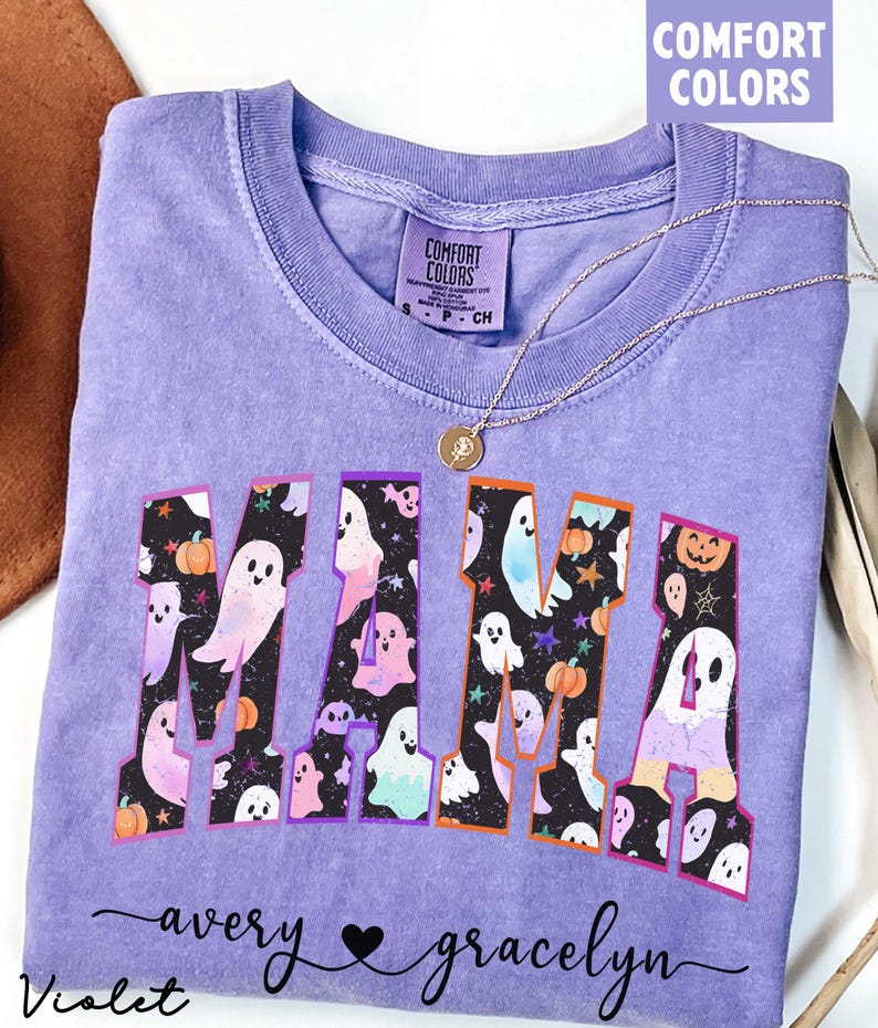 May include: A lavender Comfort Colors t-shirt with the word "MAMA" in a collegiate font, filled with ghost and pumpkin graphics. The names "avery" and "gracelyn" are written below the word "MAMA". A gold necklace with a pendant is on the shirt.